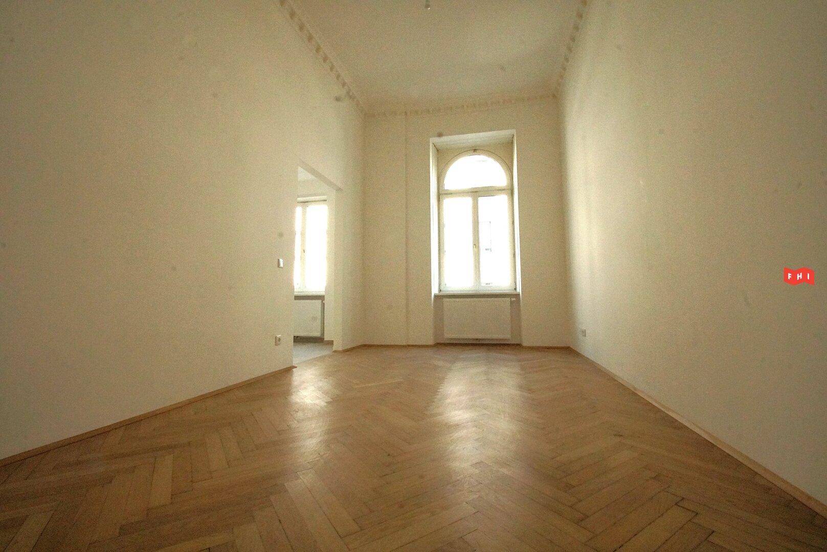 Ein geräumiger Raum mit Fischgrätparkett, einem Bogenfenster und einem Durchgang zu einem weiteren Zimmer.