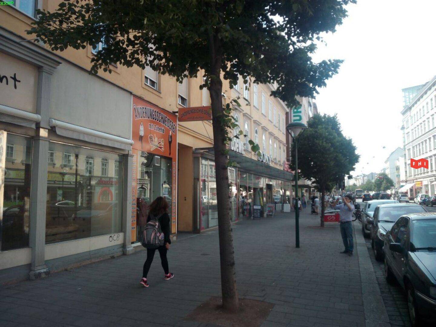 Blick auf eine belebte Stadtstraße mit Geschäften im Erdgeschoss, parkenden Autos und Bäumen entlang des Bürgersteigs.