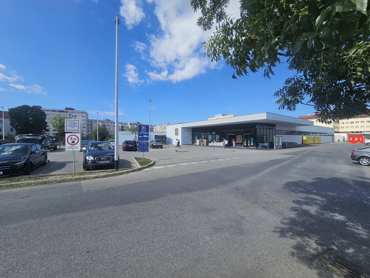 Außenansicht eines Supermarktes mit großem Parkplatz unter blauem Himmel und umliegenden Gebäuden.
