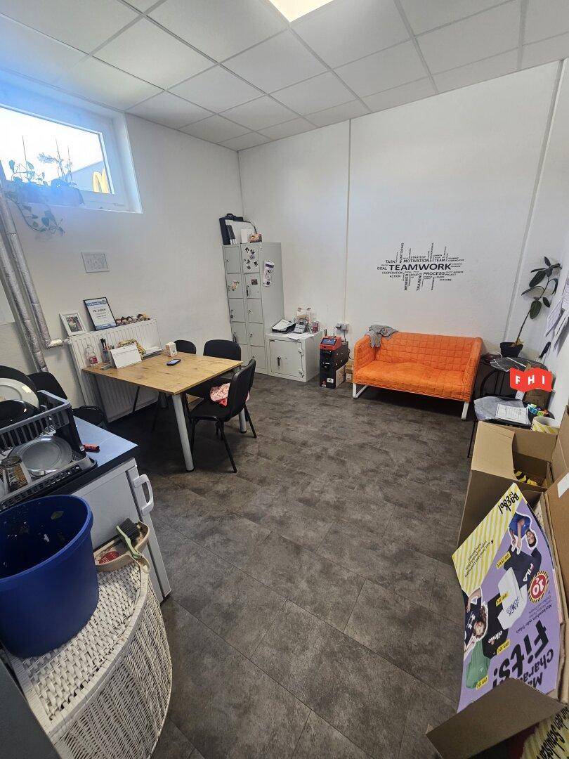 Kleines Büro oder Pausenraum mit Tisch, Sofa und Schränken, gut beleuchtet durch ein Fenster.
