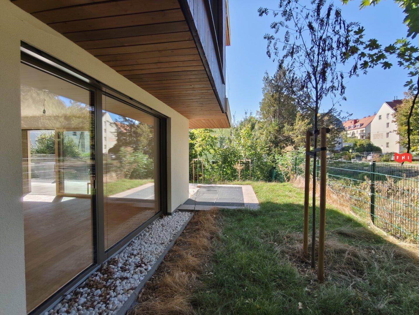 Kleiner Gartenstreifen neben dem Gebäude mit großen Fenstern und Blick ins Grüne.