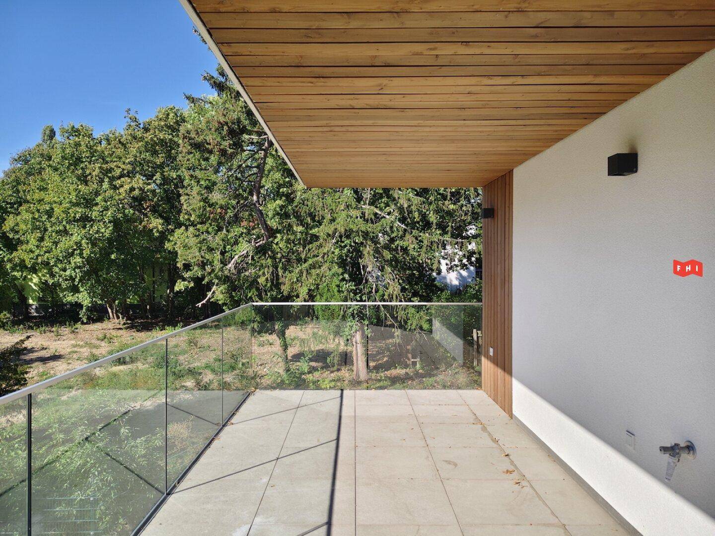 Heller Balkon mit Fliesenboden und Glasgeländer, umgeben von viel Grün.