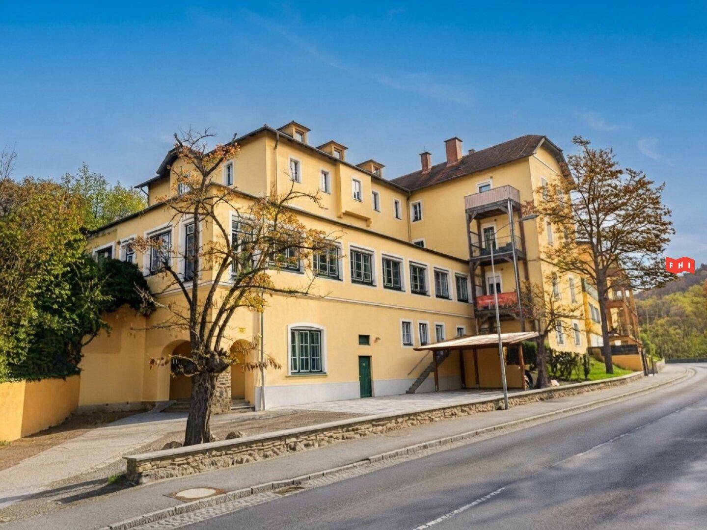 Historisches Gebäude mit gelber Fassade, grünen Fensterläden und Blick auf eine bewaldete Anhöhe.