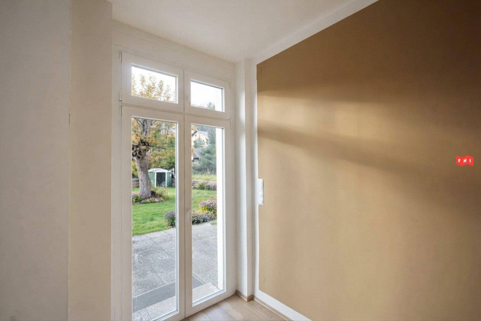 Heller Innenraum mit beiger Wand und einer Glastür, die Zugang zu einem grünen Gartenbereich bietet.