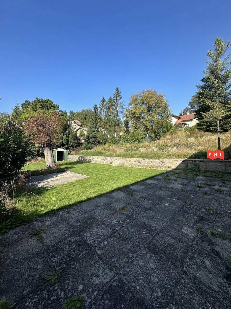 Gepflegter Gartenbereich mit Rasenfläche, gepflasterter Terrasse und umgebenden Bäumen unter blauem Himmel.