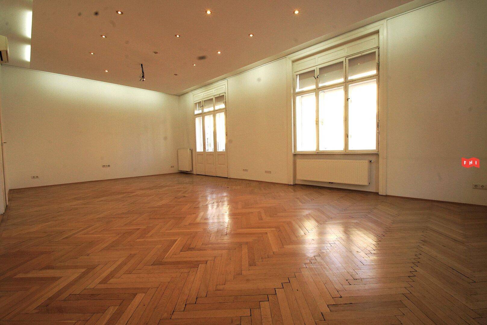 Heller, leerer Raum mit Fischgrätparkett, großen Fenstern und Einbaustrahlern in der Decke.