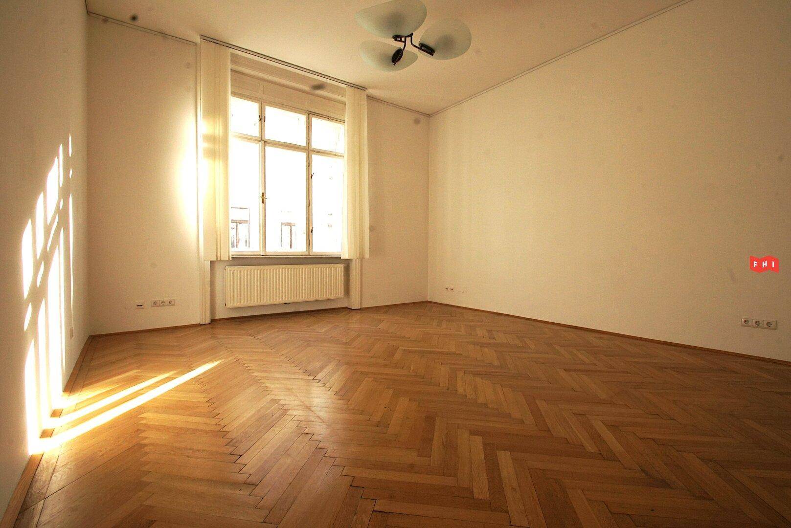 Heller Raum mit Fischgrätparkett, einem großen Fenster und Heizkörper, durchflutet von Sonnenlicht.