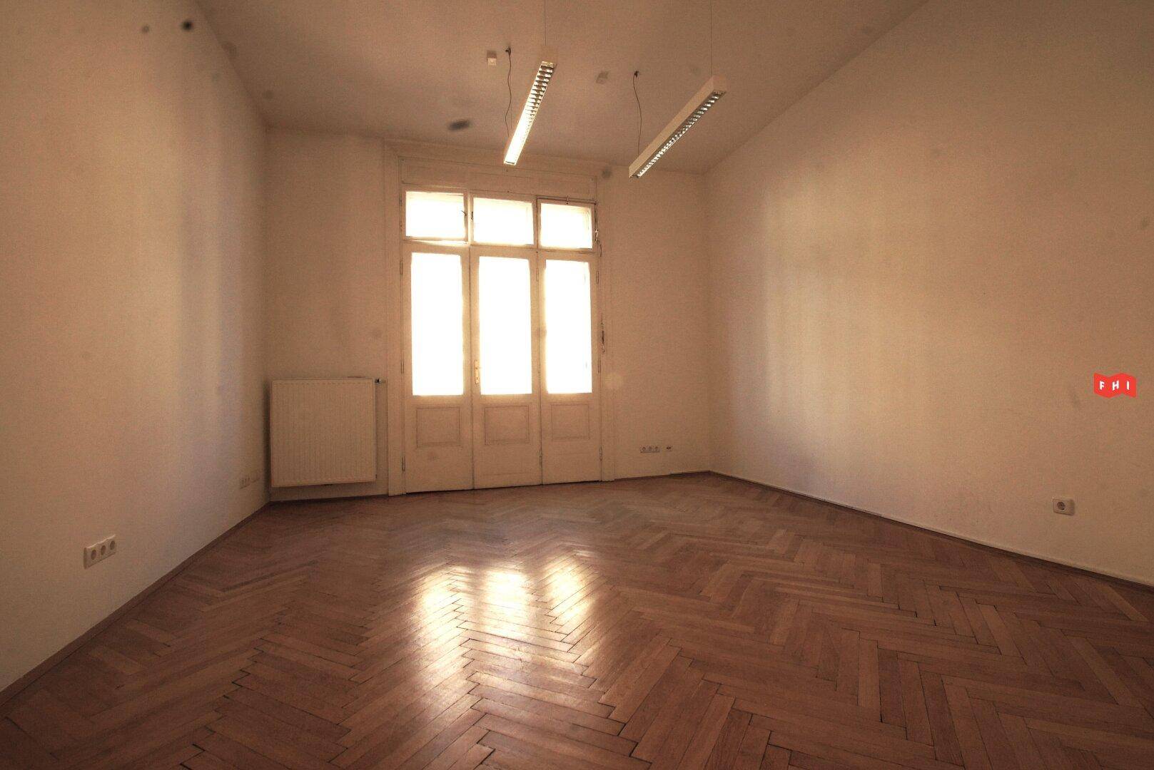 Heller, leerer Raum mit Fischgrätparkett, Deckenleuchten und einem großen Fenster mit Zugang nach draußen.