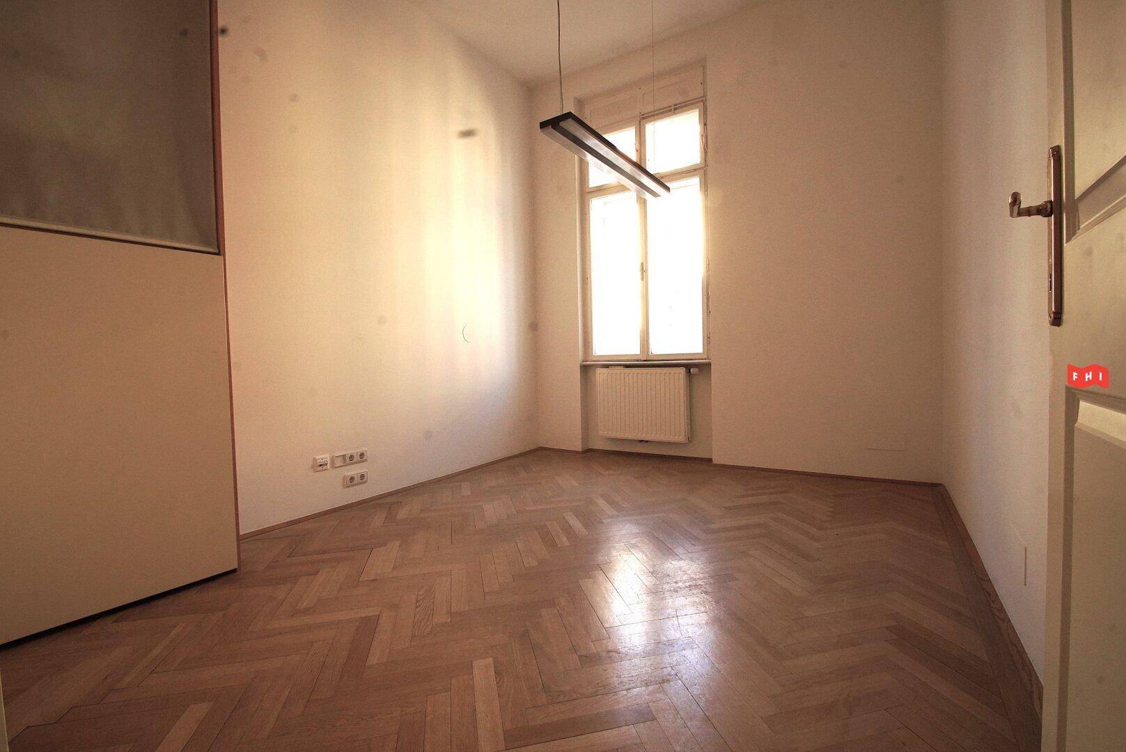 Heller, leerer Raum mit Fischgrätparkett, einem Fenster und einer modernen Hängelampe.