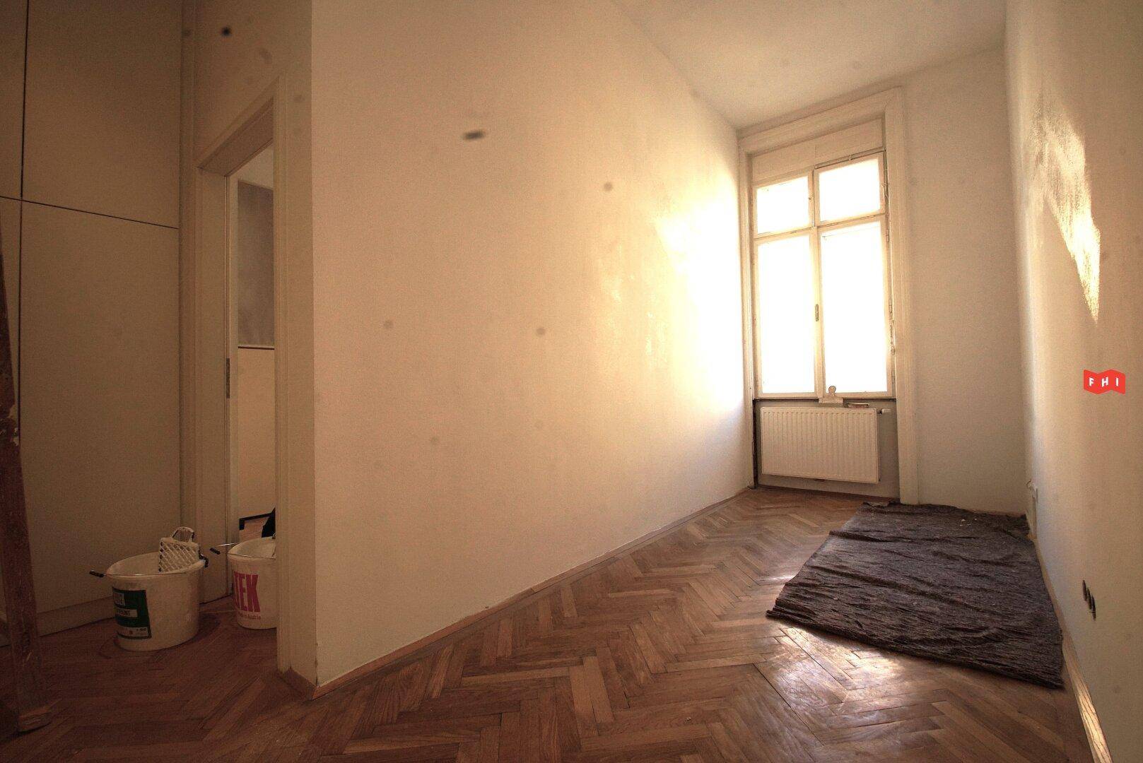 Raum mit Fischgrätparkett und Fenster, angrenzend an einen weiteren Raum, möglicherweise in Vorbereitung für Renovierung.