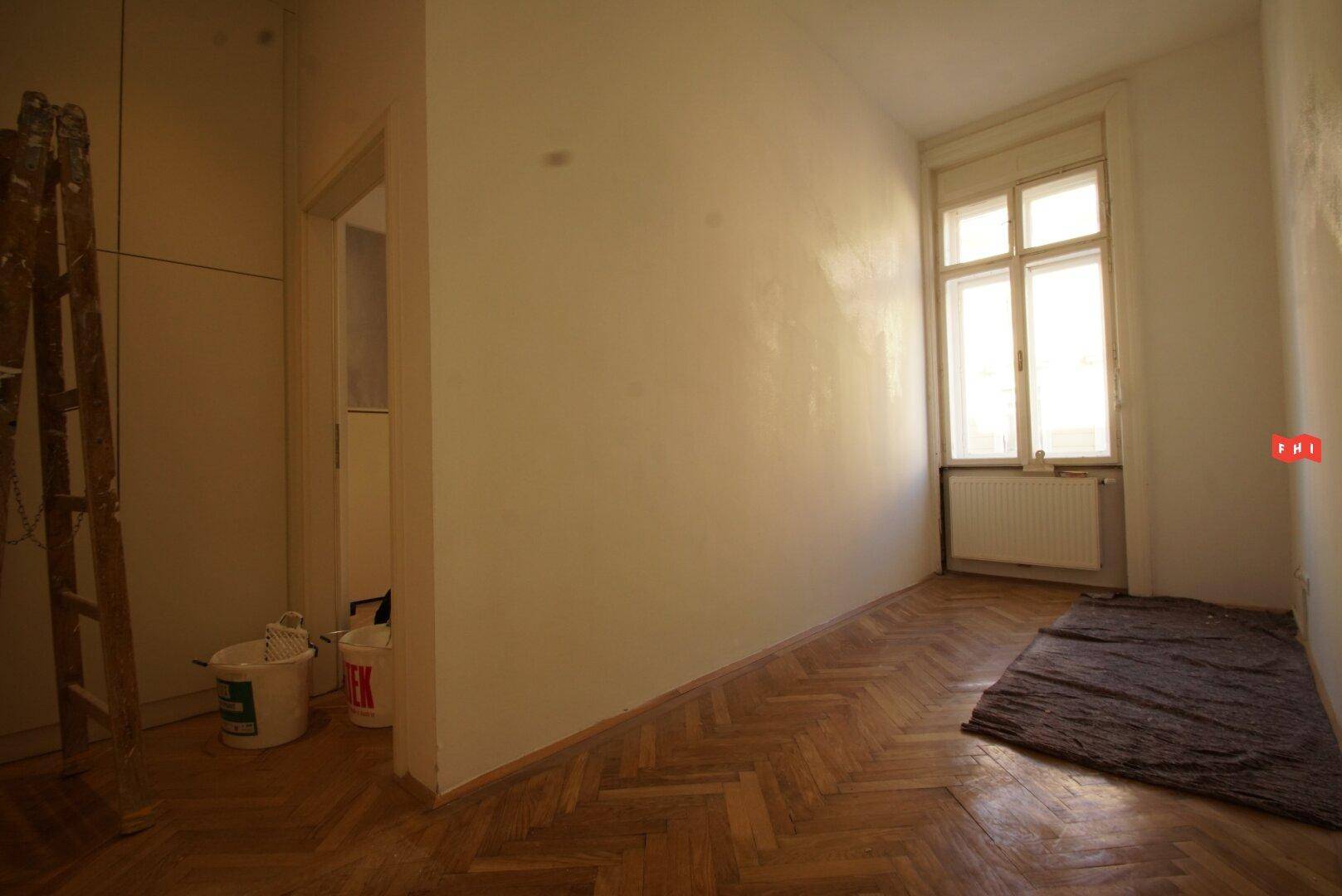 Raum mit Fischgrätparkett und Fenster, angrenzend an einen weiteren Raum, mit Leiter und Eimern.