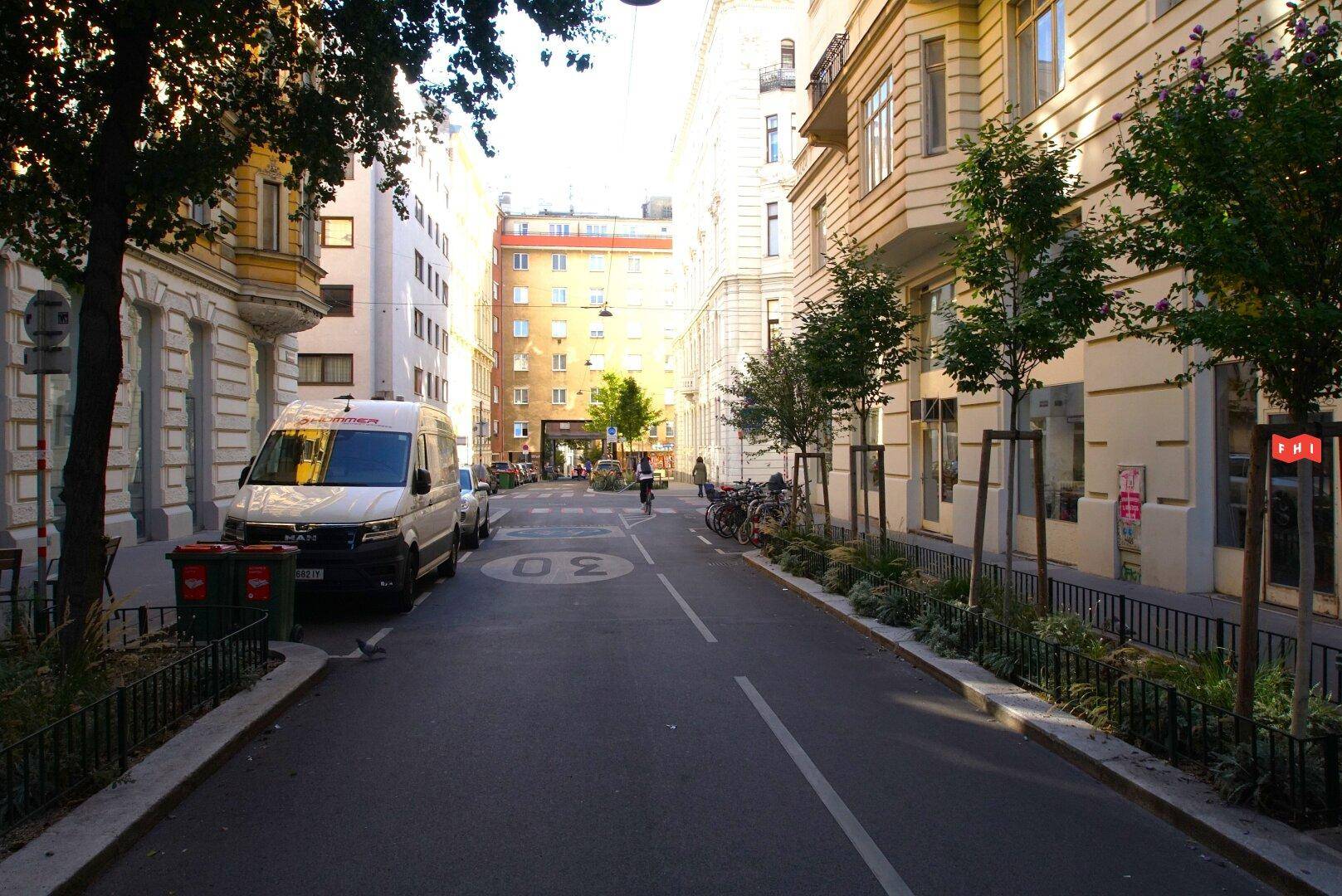 Blick auf eine ruhige Stadtstraße mit historischen Gebäuden, geparkten Fahrzeugen und Bäumen.