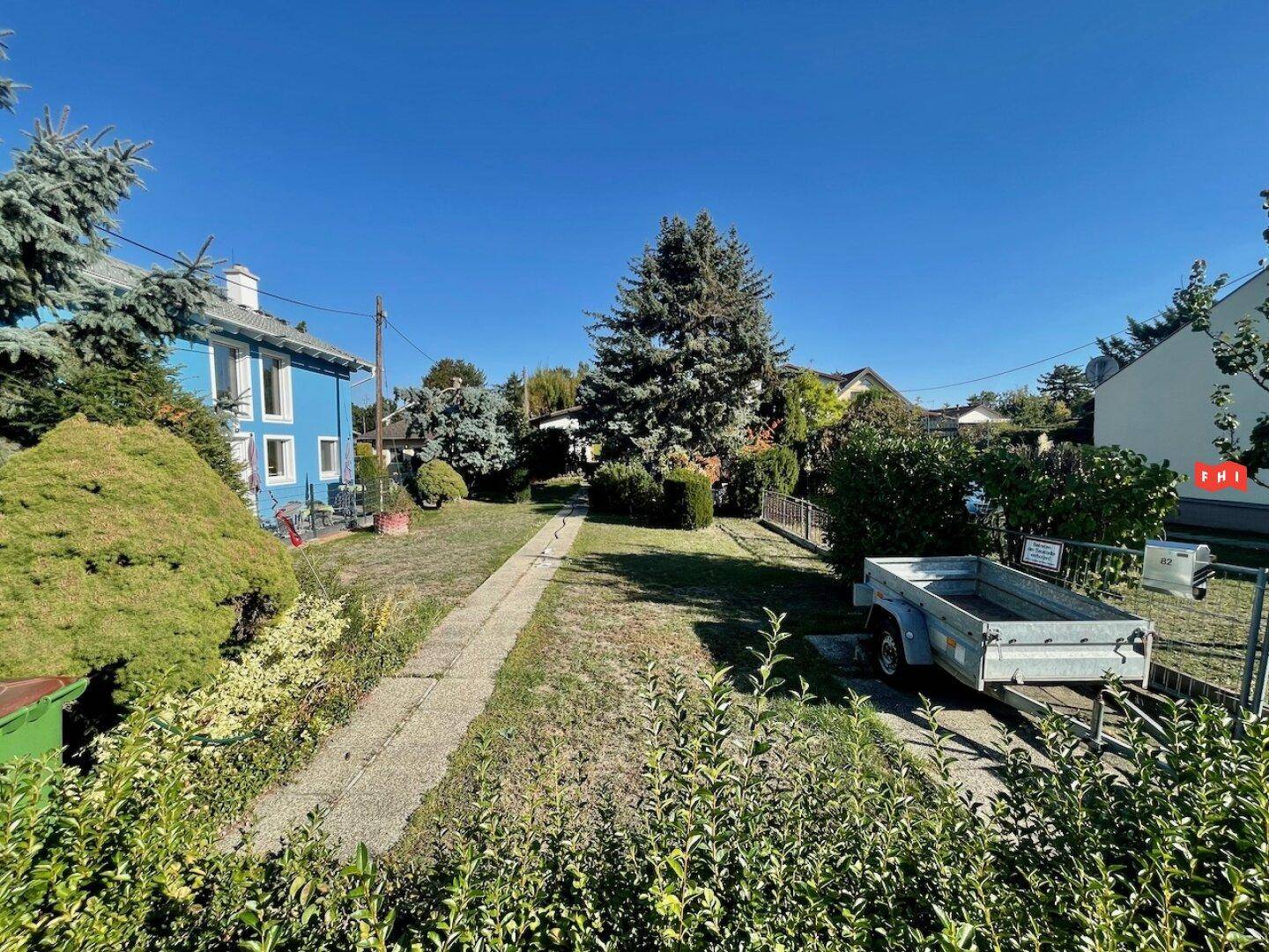 Weitläufiger Garten mit einem gepflasterten Weg, umgeben von Bäumen und einem blauen Haus im Hintergrund.