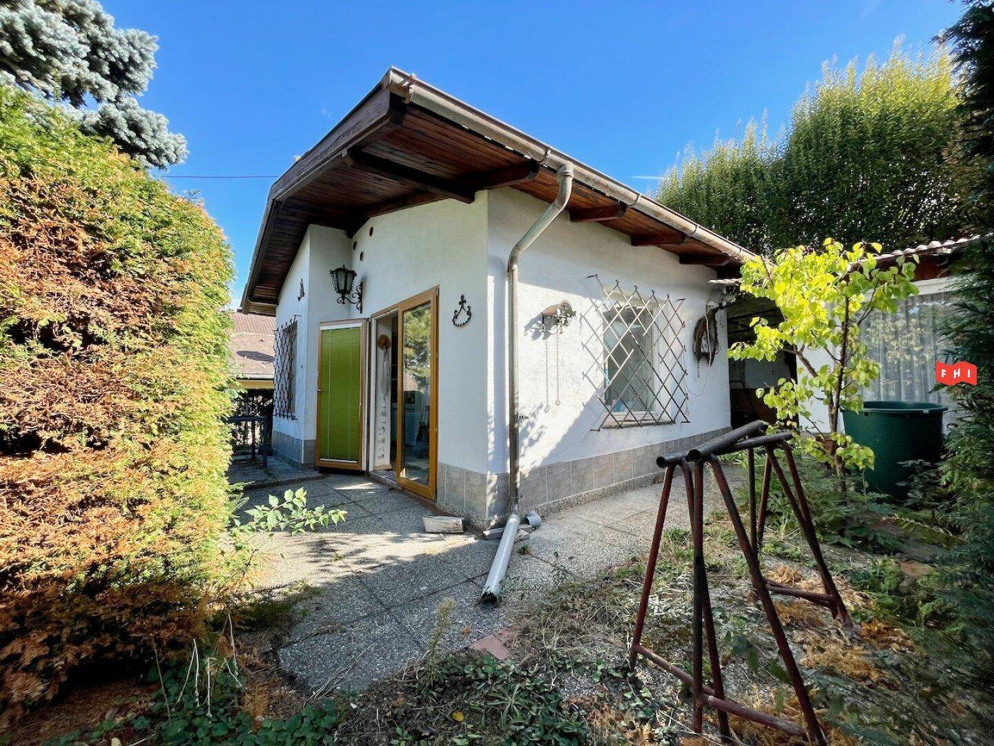 Kleine Hütte mit weißer Fassade und Holzdach, umgeben von Grün, mit einem gepflasterten Terrassenbereich.