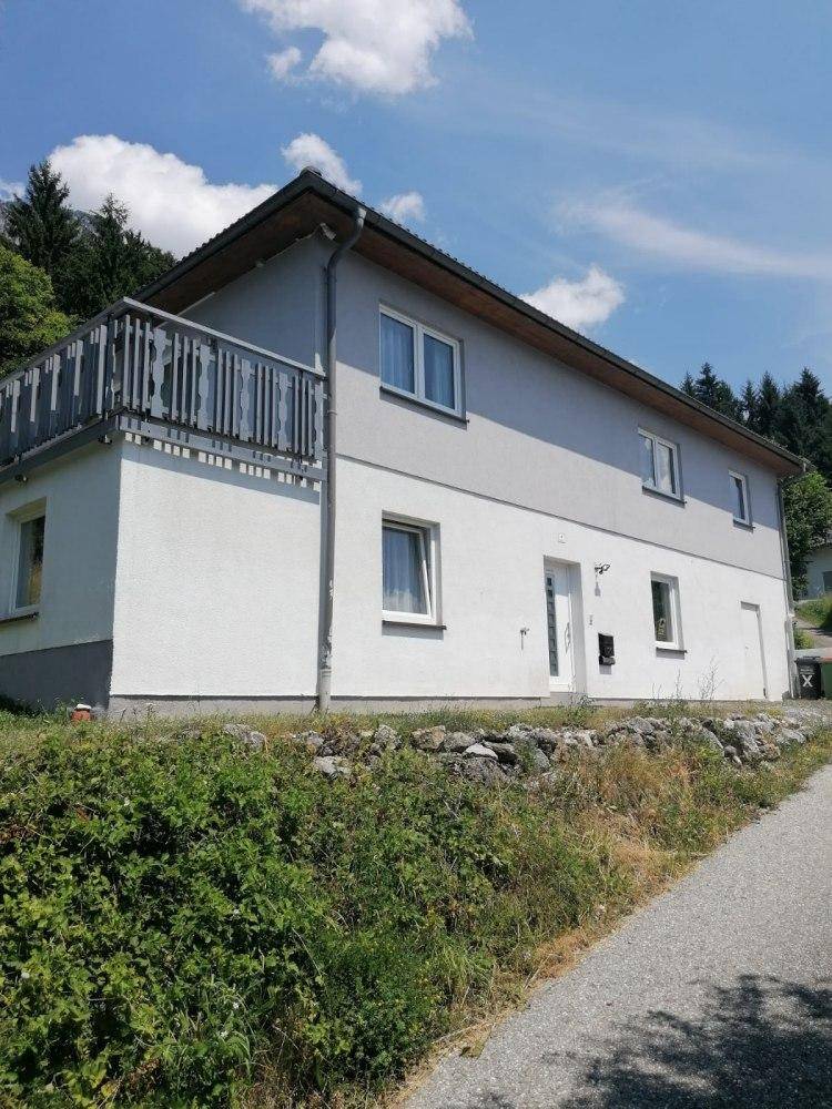 Seitenansicht des Hauses mit Balkon und grüner Umgebung unter blauem Himmel.