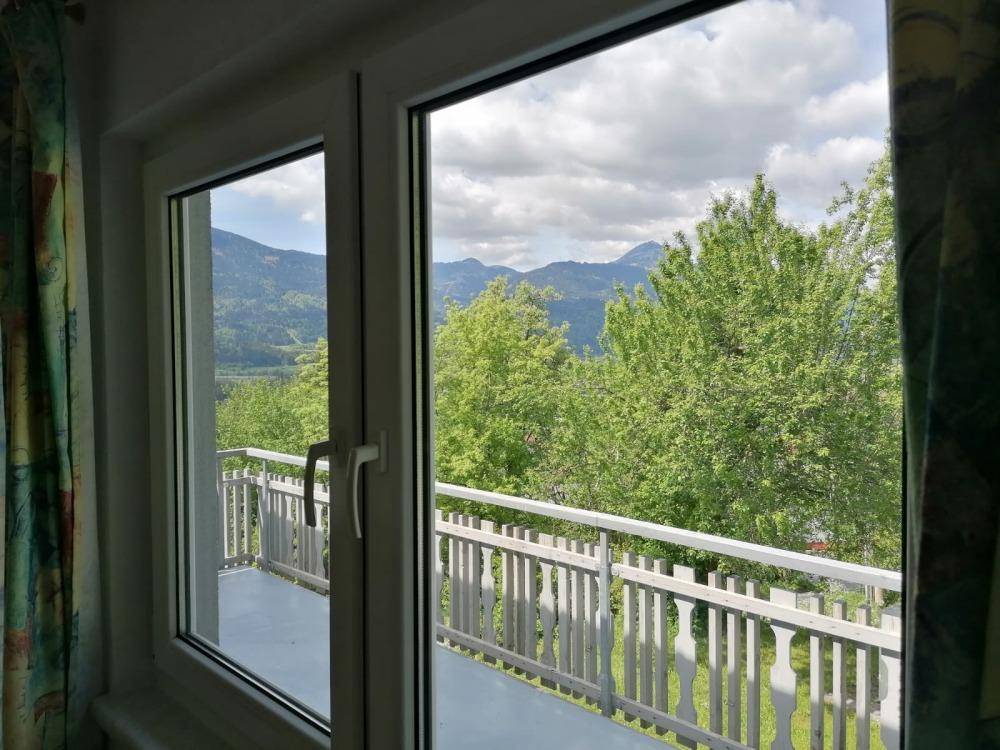 Blick aus dem Fenster auf den Balkon mit Holzbalkongeländer und die grüne Berglandschaft.