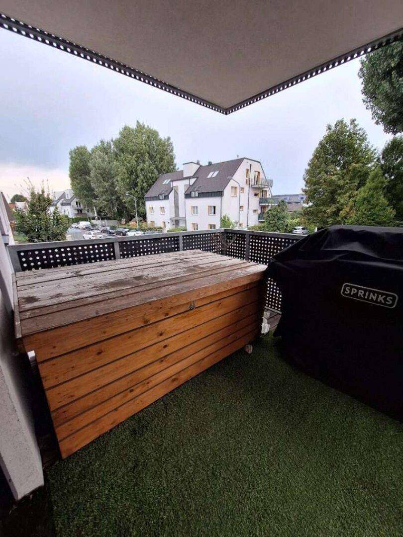Balkon mit Kunstrasen, Holzbank und Grill, umgeben von Bäumen.