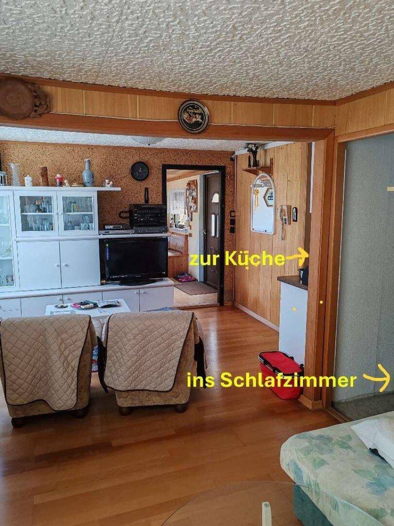 Wohnzimmer mit Holzvertäfelung, Fernseher und Durchgängen zur Küche und zum Schlafzimmer.