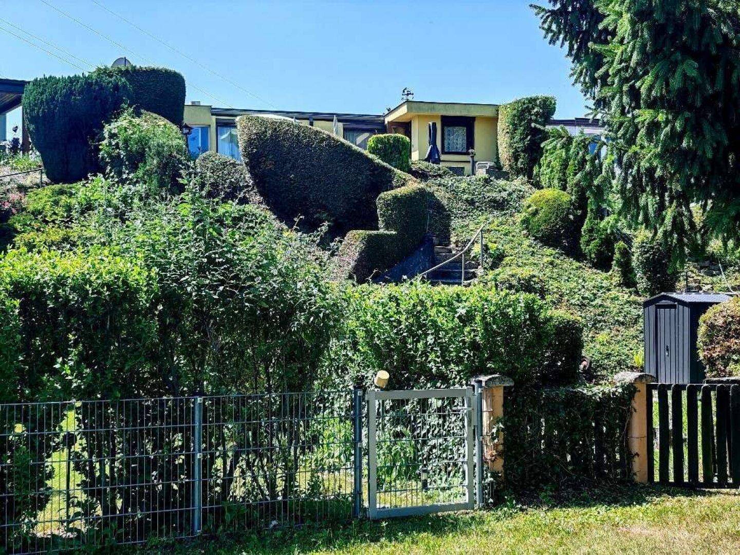 Gepflegter Gartenbereich mit Hecken, Sträuchern und einem Gartenhaus auf einem Hanggrundstück.