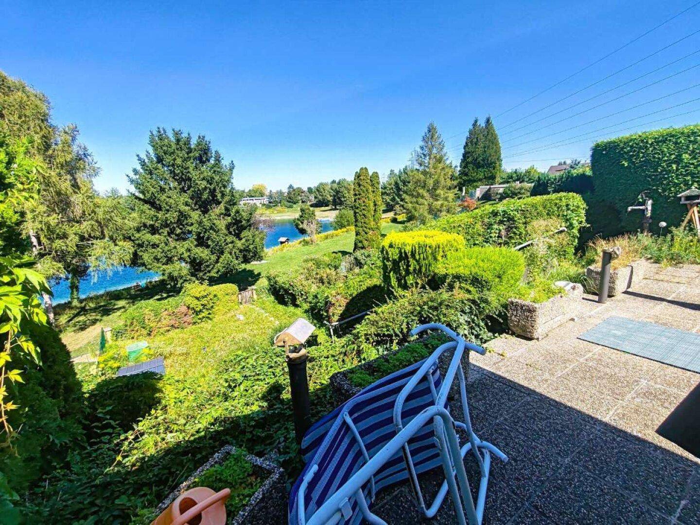 Terrasse mit Blick auf den weitläufigen Garten und den See, umgeben von grüner Natur.