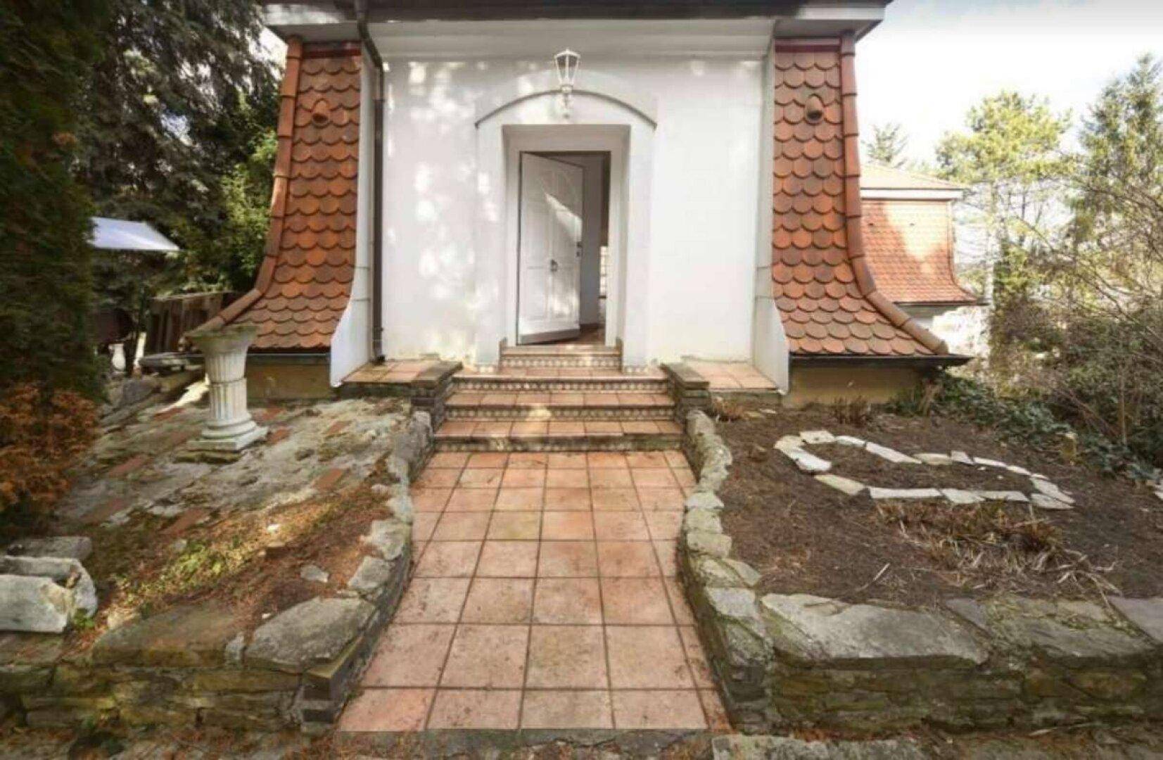 Eingangsbereich des Hauses mit gefliestem Weg, Steinstufen und dekorativen Elementen im Garten.