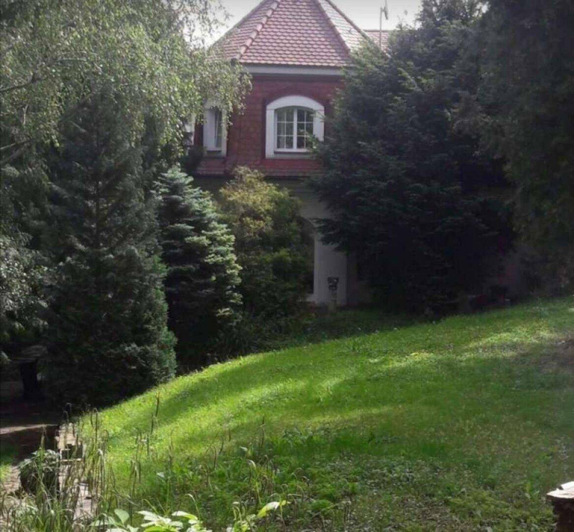 Rotes Haus mit Mansarddach, umgeben von hohen Bäumen und üppigem Grün.