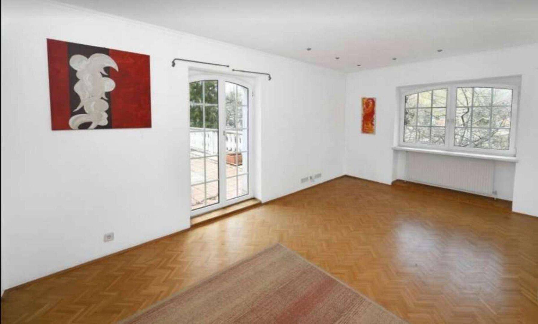 Helles Wohnzimmer mit Fischgrätparkett, großen Fenstern und Zugang zu einem Balkon.