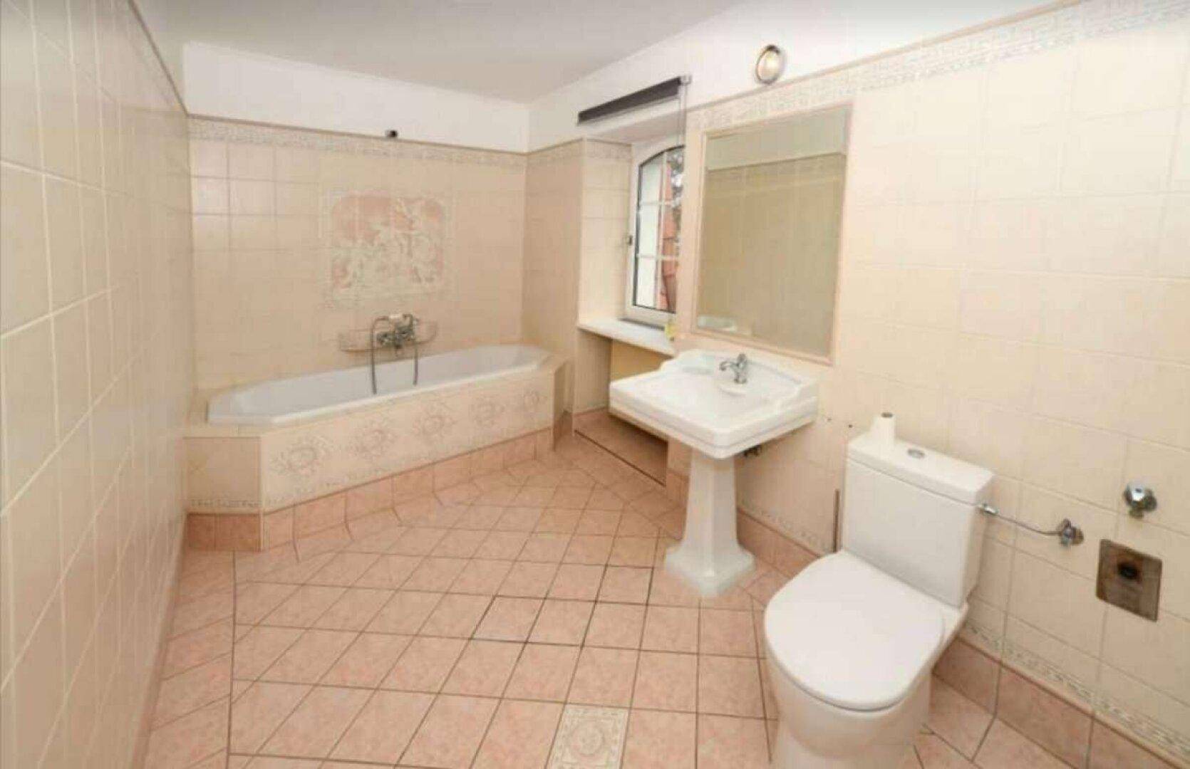 Badezimmer im 80er-Jahre-Stil mit beigefarbenen Fliesen, Badewanne und Fenster.
