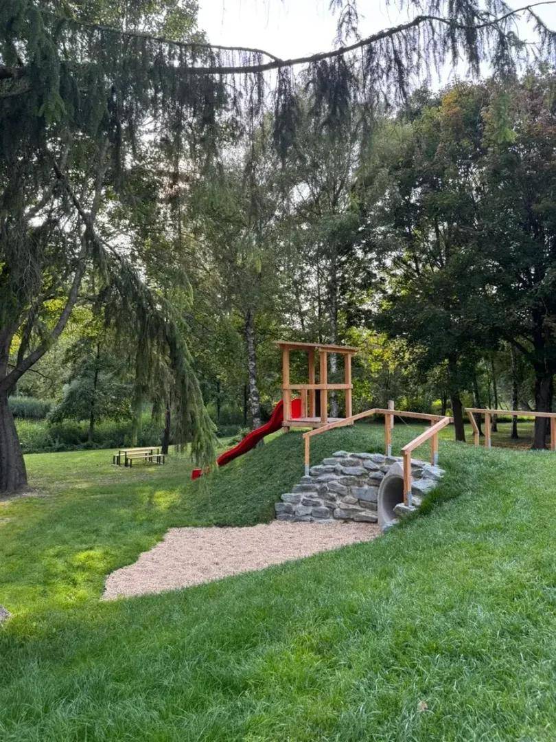 Naturnaher Spielplatz mit Rutsche, Klettergerüst und Tunnel in einer grünen Parklandschaft.