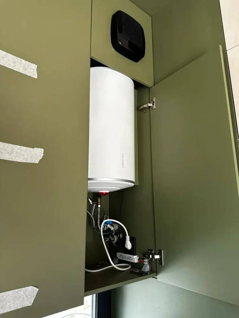 Warmwasserbereiter in einem olivgrünen Schrank, der die technische Ausstattung des Raumes zeigt.