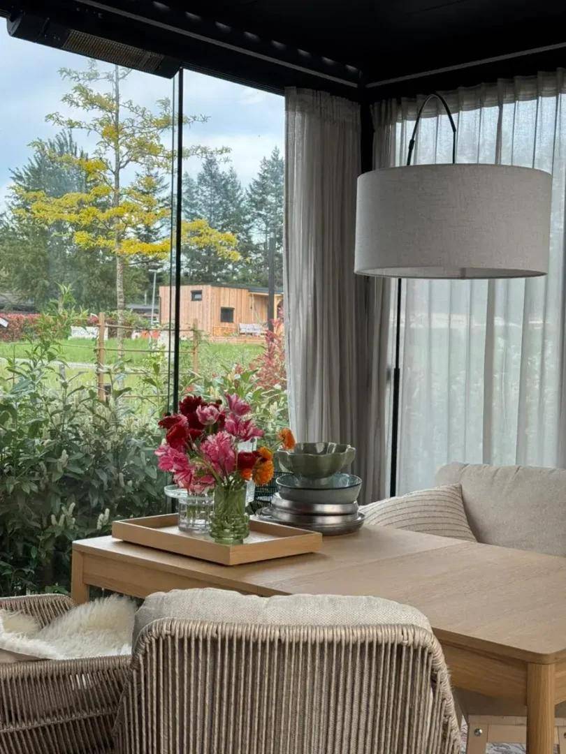 Heller Essbereich mit Holztisch und Korbstühlen, umgeben von großen Fenstern mit Gartenblick.