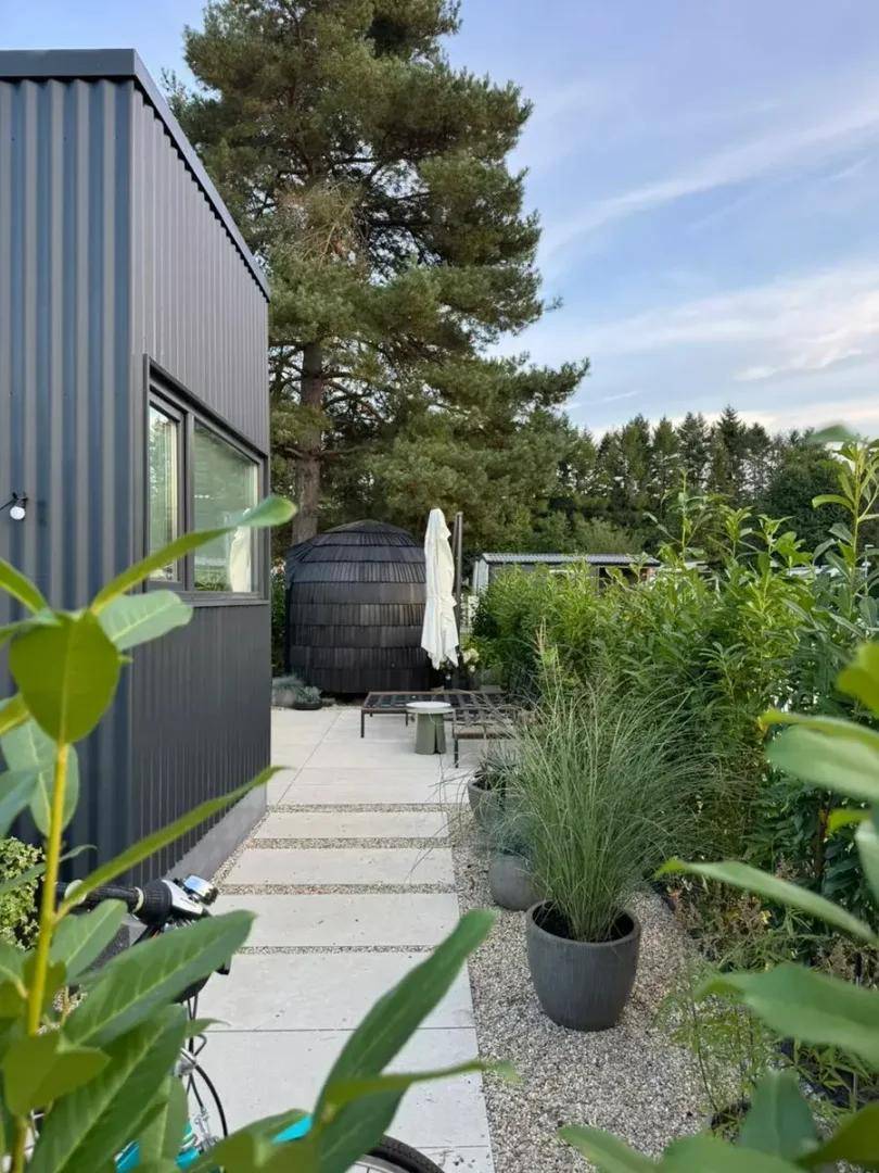 Gepflasterte Terrasse mit einem Saunafass, Liegestühlen und üppiger Bepflanzung im Gartenbereich.