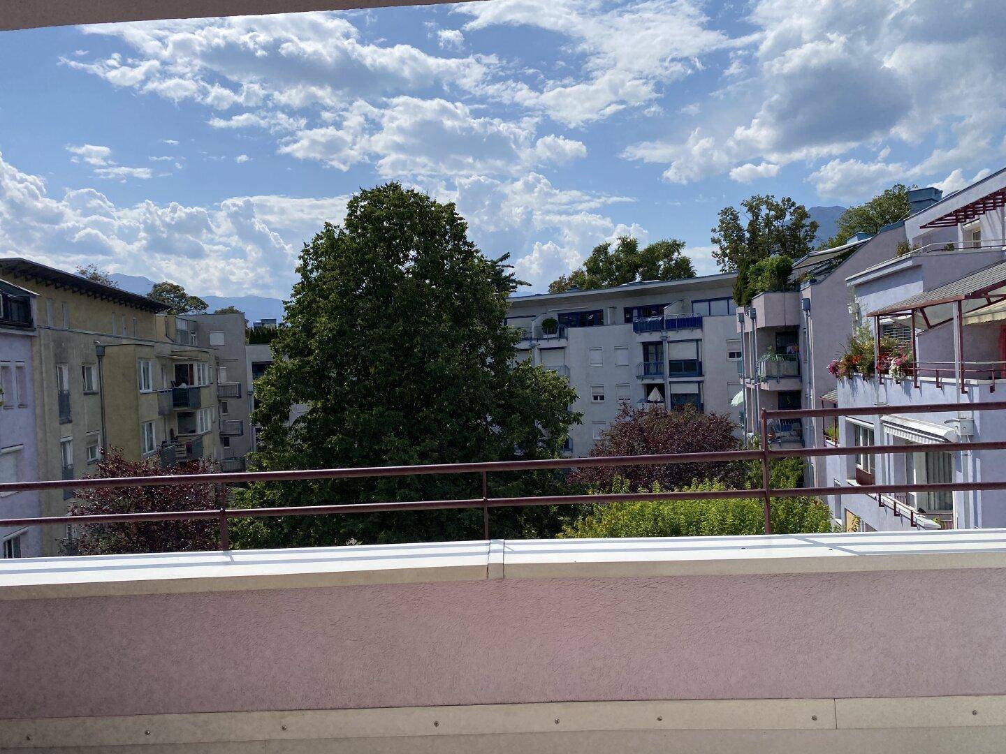 Balkon mit weitem Blick über die Stadt, grüne Bäume und ferne Berge.