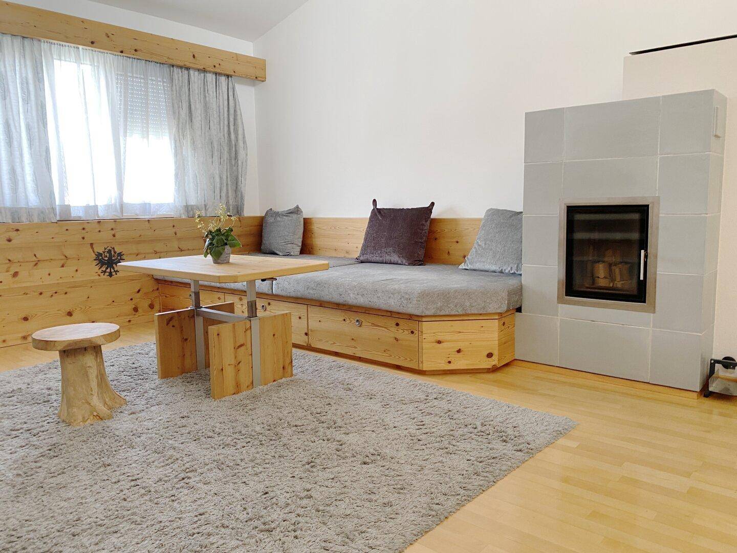 Einladendes Wohnzimmer mit Kachelofen, Holzbänken und einem hellen Teppich.