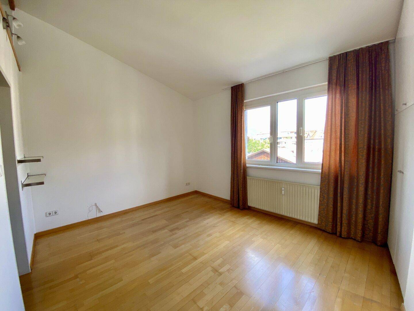 Helles Schlafzimmer mit Holzparkett und einem großen Fenster mit braunen Vorhängen.
