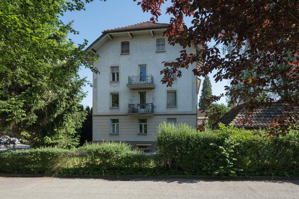 Fassade eines Altbaus mit kleinen Balkonen, umgeben von Bäumen und Sträuchern.