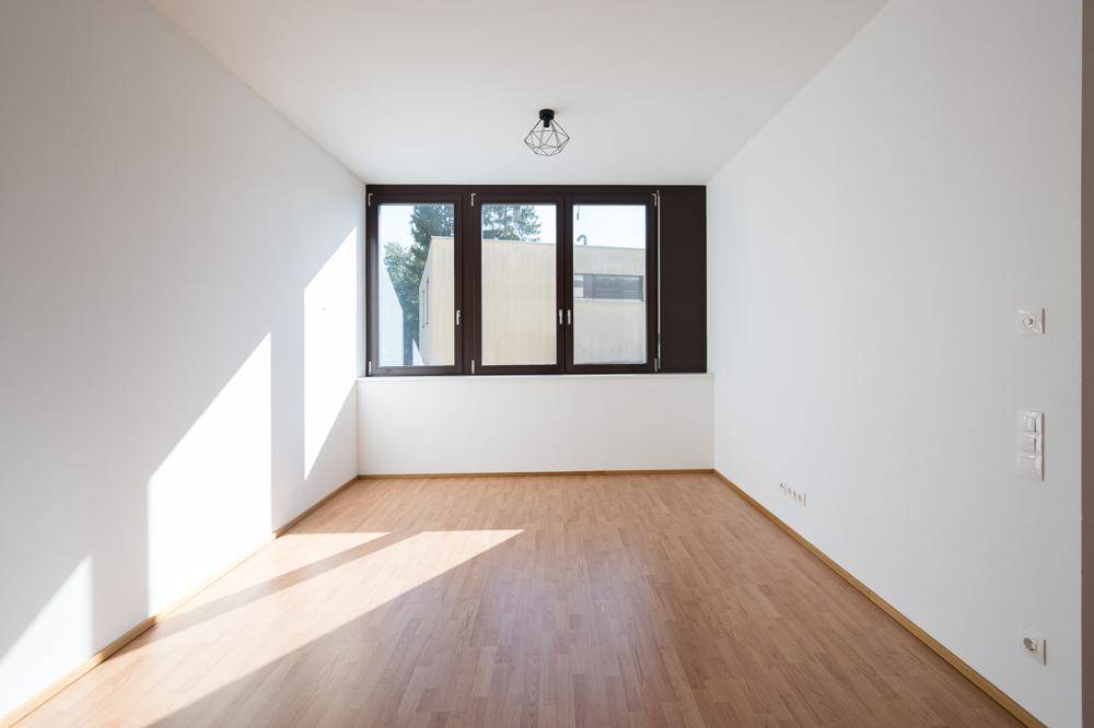 Heller, leerer Raum mit großen Fenstern und Laminatboden, durchflutet von natürlichem Sonnenlicht.
