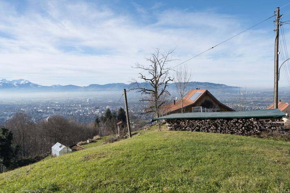 Weitläufiger Ausblick über das Tal mit Stadt, Bergen und einem Haus mit Holzstapel.