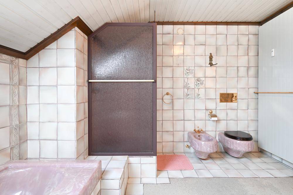 Badezimmer mit rosa Bidet, Toilette, Duschbereich und weißen Fliesen an Wänden und Boden.