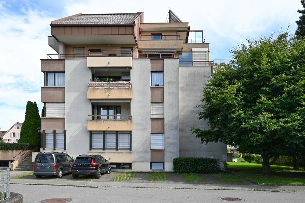 Mehrstöckiges Wohngebäude mit Balkonen, großen Fenstern und Parkplätzen davor.