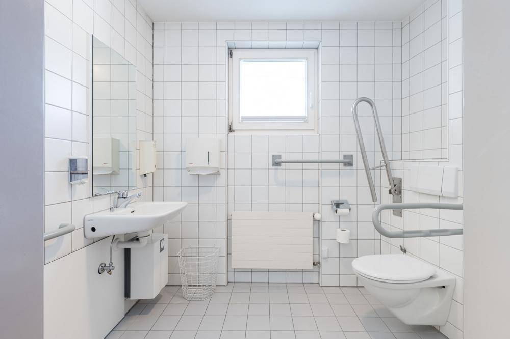 Helles, gefliestes Badezimmer mit Fenster, Toilette, Waschbecken und Haltegriffen für Barrierefreiheit.