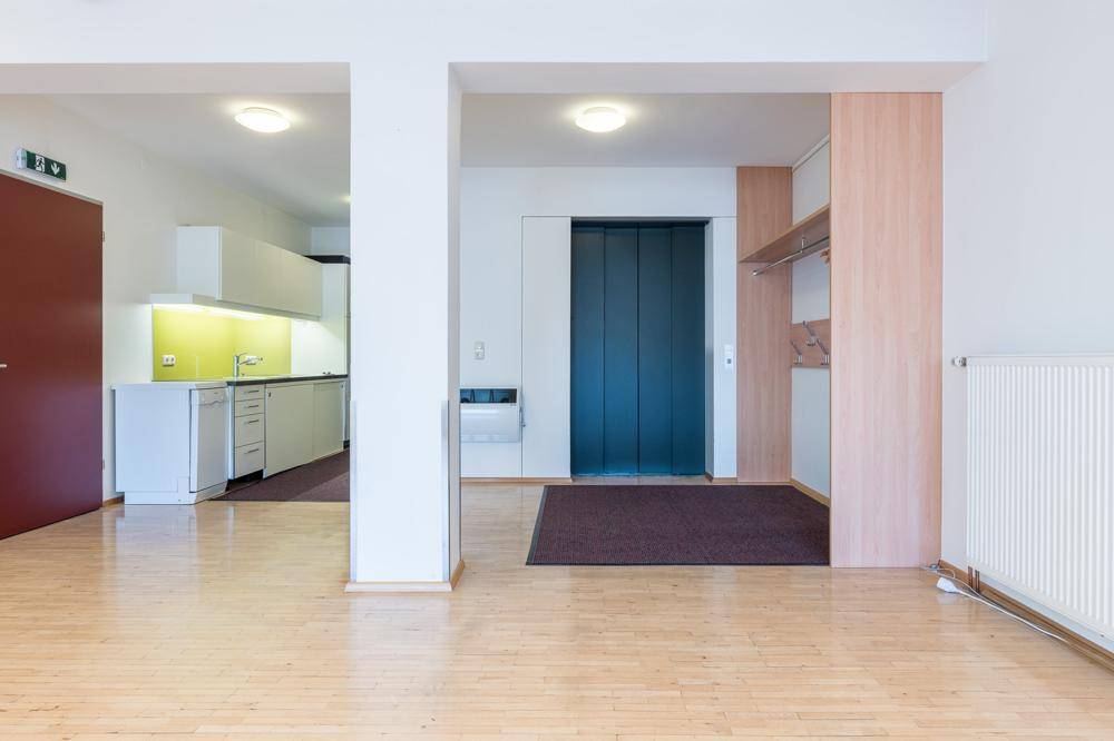 Offener Gemeinschaftsbereich mit Holzboden, integrierter Küchenzeile, Aufzugzugang und Garderobenbereich.