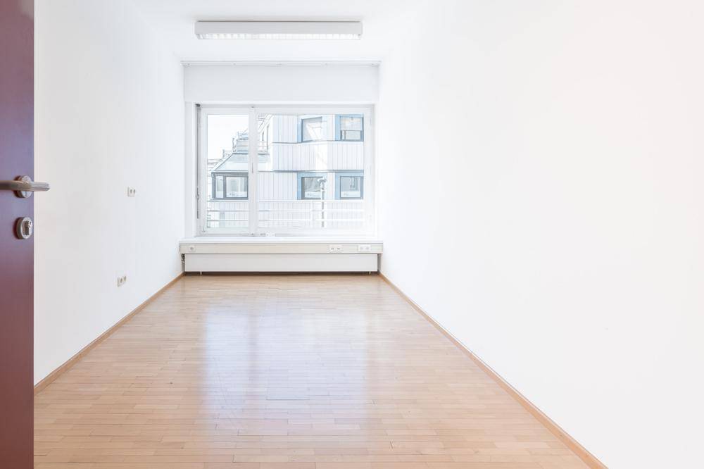 Schmaler, heller Raum mit Holzboden und einem Fenster, das einen Blick auf die umliegenden Gebäude freigibt.