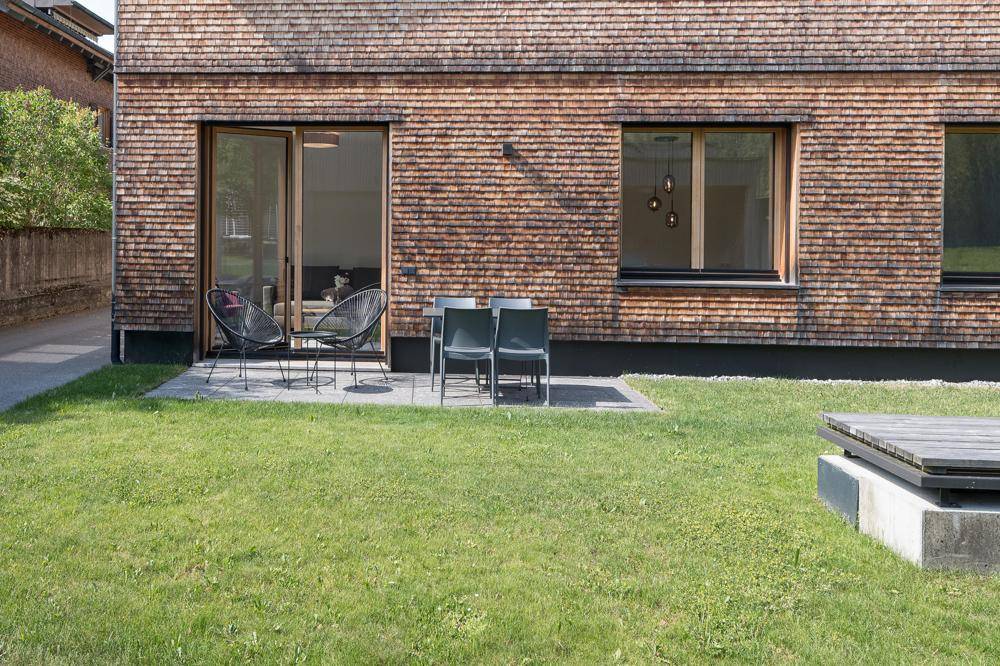 Rückseite des Hauses mit Holzschindelfassade, Rasenfläche und einer möblierten Terrasse.