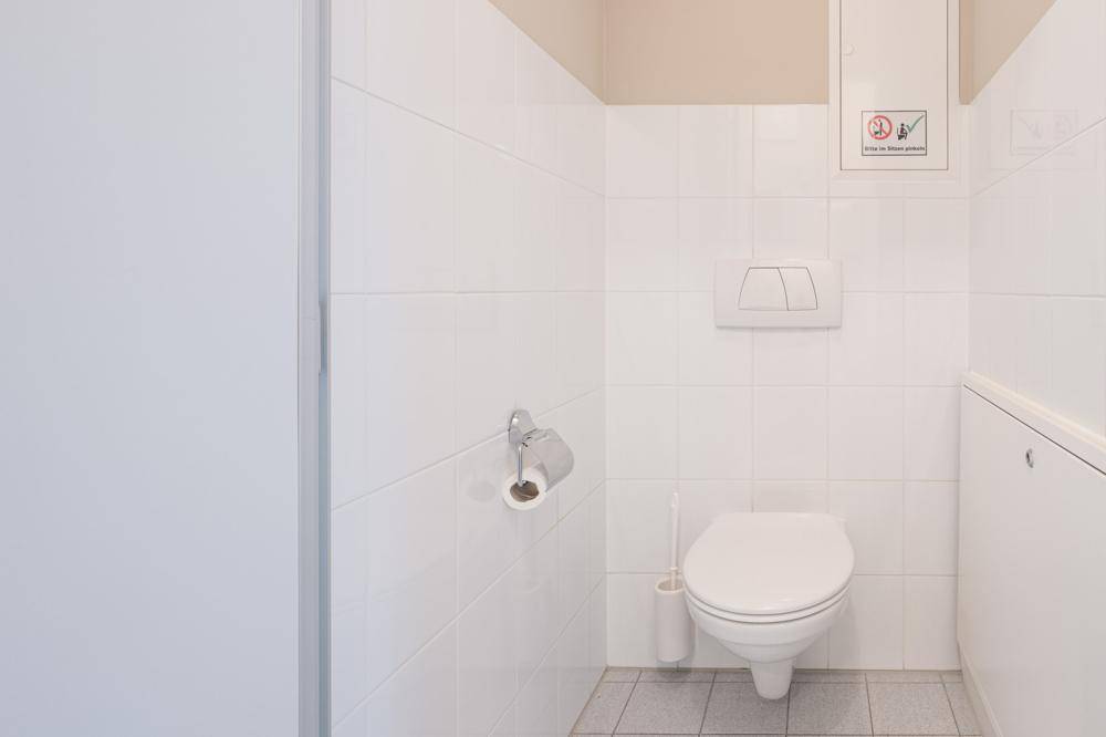 Saubere Gästetoilette mit Wand-WC und weißen Fliesen, ausgestattet mit Toilettenpapierhalter.