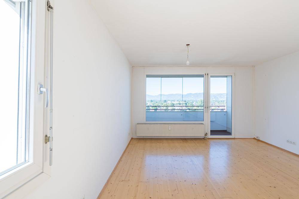 Helles, leeres Zimmer mit Holzparkettboden und Zugang zu einem verglasten Balkon mit Bergblick.