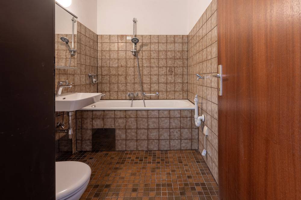Badezimmer mit braunen Fliesen, Badewanne mit Duschfunktion und einem einzelnen Waschbecken.