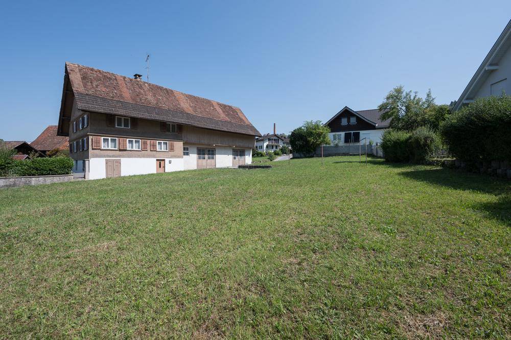 Sonniger Gartenbereich mit Rasen und einem traditionellen Haus im Hintergrund, umgeben von Natur.