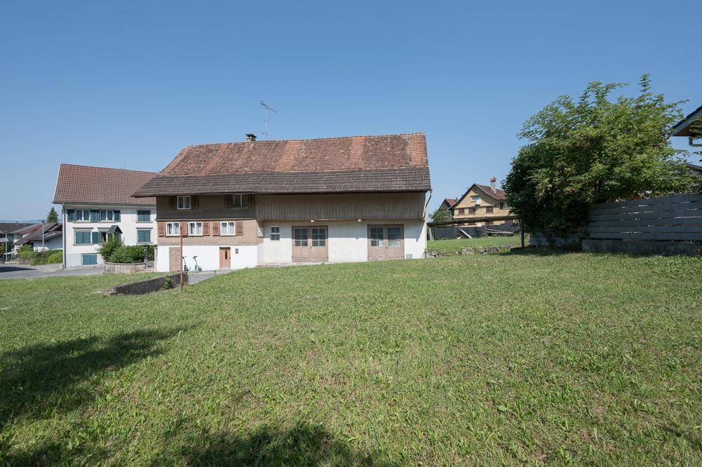 Offene Rasenfläche vor einem traditionellen Gebäude, umgeben von weiteren Häusern und viel Grün.