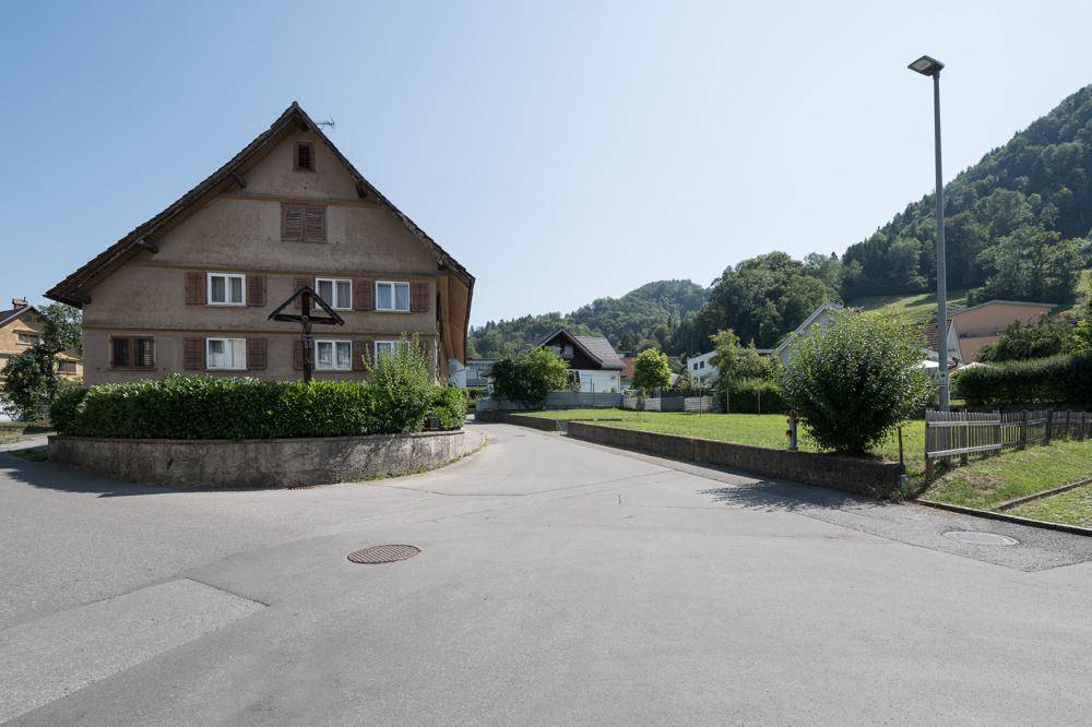 Traditionelles Haus an einer ländlichen Straße mit einem Holzkreuz und bewaldeten Hügeln im Hintergrund.